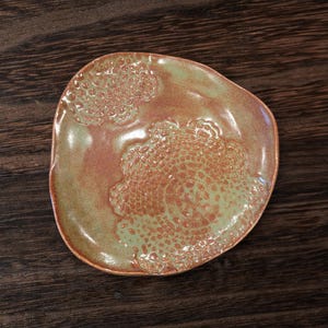 May include: A handmade ceramic plate with an irregular shape. The plate has a textured, lace-like design in green and reddish-brown hues. This decorative plate is suitable for serving food or displaying small items, and is a unique piece.