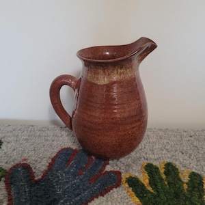 Puede incluir: Una jarra de cerámica hecha a mano, de color marrón rojizo, con un asa curva y un pico. La jarra tiene una textura moteada y un borde de color más claro. Es una pieza rústica y artesanal.