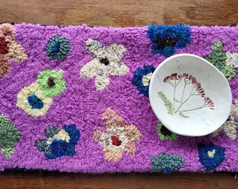 以下が含まれることがあります： 青、緑、クリーム色など、さまざまな色の刺繍の花柄が施された、手作りの紫色のラグ。植物のイラストが描かれた白い楕円形のボウルがラグの上に置かれています。手作り。