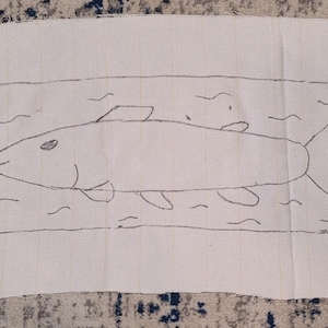 May include: A rectangular, off-white fabric with a hand-drawn illustration of a fish swimming in water. The fish is outlined in black, with details like fins and an eye. The fabric has a simple, artistic design.