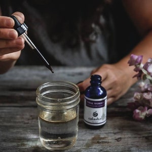 Op de afbeelding: Een persoon voegt druppels vloeistof toe vanuit een druppelfles in een glazen pot. De pot is gevuld met een heldere vloeistof. Twee flessen met etiketten waarop "Vital Women" en "Peaceful Mind" staat, zijn zichtbaar op een houten oppervlak. De flessen zijn blauw en hebben een wit etiket met zwarte tekst. De flessen zijn van het merk "Calasha".