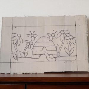 May include: A rectangular piece of beige fabric with a hand-drawn design of a beehive, flowers, and a small bird. The design is outlined in black. The dimensions are 17 x 9.5 inches.