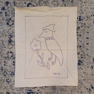 May include: A cream-colored fabric panel featuring a hand-stitched bird design in purple thread. The bird wears a hat and holds a flower. The dimensions are 19 x 13 inches.