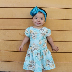 May include: A young girl wearing a light blue floral dress with a turquoise bow headband. She is standing in front of a wood panel wall and smiling at the camera.