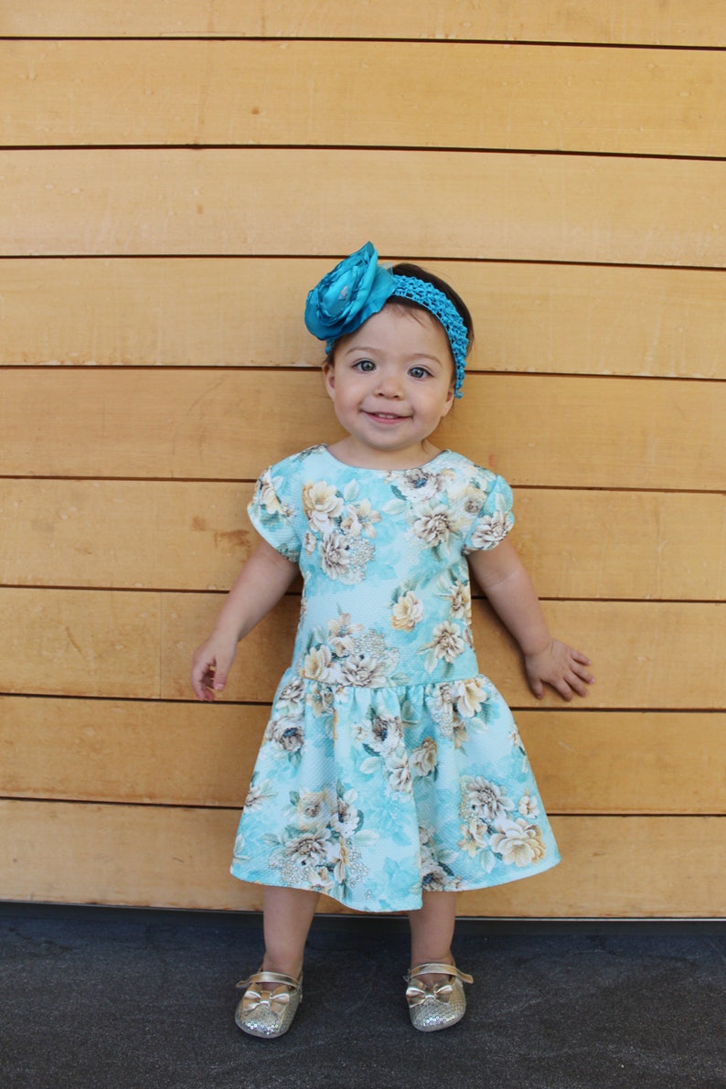 May include: A young girl wearing a light blue floral dress with a turquoise bow headband. She is standing in front of a wood panel wall and smiling at the camera.