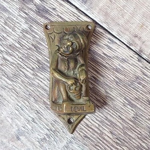 May include: A brass door knocker shaped like a devil holding a skull. The knocker is mounted on a wooden surface and has the word "Devil" inscribed on it.