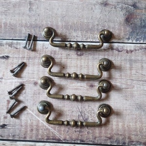 以下が含まれることがあります： 装飾的なビーズと丸みを帯びた端を持つ、アンティークブラスのキャビネットプル4個。プルは、横にネジが付いた状態で一列に並んでいます。