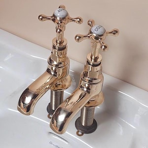 May include: Two shiny gold-colored faucets with cross-shaped handles labeled "HOT" and "COLD" are mounted on a white porcelain sink. The faucets have a classic design and are positioned against a light peach-colored wall.