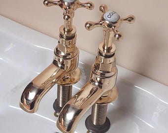 Puede incluir: Dos grifos dorados brillantes con manijas en forma de cruz, marcados "HOT" y "COLD", están montados en un lavabo de porcelana blanca. Los grifos tienen un diseño clásico y están colocados contra una pared de color melocotón claro.