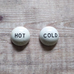 May include: Two white ceramic knobs with black lettering. The left knob says "HOT" and the right knob says "COLD".
