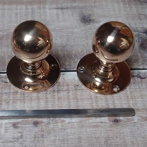 May include: Two polished copper door knobs with round handles and a circular base. The knobs are on a wooden surface.