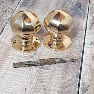 Peut inclure: Deux boutons de porte dorés avec un design facetté et une base carrée. Les boutons sont sur une surface en bois avec une vis au centre de l'image.