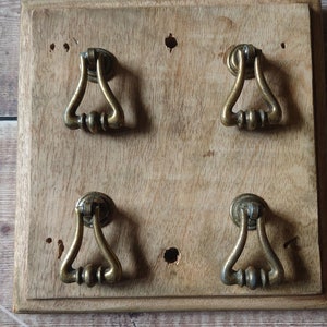 May include: Four antique brass cabinet door pulls mounted on a wooden board. The pulls have a teardrop shape with a decorative ring around the center.