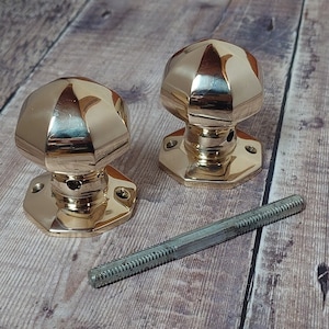 May include: Two polished brass door knobs with a faceted design. The knobs are on a wooden surface with a metal screw.
