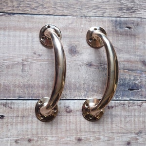 May include: Two polished brass door handles with a curved, arching design. Each handle features two circular mounting plates at the ends, each containing three pre-drilled screw holes for installation. The metal has a smooth, reflective finish.