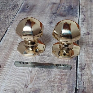 May include: Two gold door knobs with a square base and a rounded top. The knobs are on a wooden surface with two small screws and a metal bar.