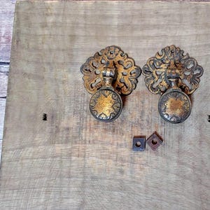 Peut inclure: Deux poignées de tiroir en métal doré orné avec des motifs complexes. Les poignées ont un bouton rond et un cadre décoratif.