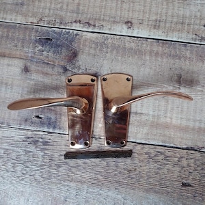 May include: Two antique copper door handles with a curved lever design. The handles are on a wooden surface.