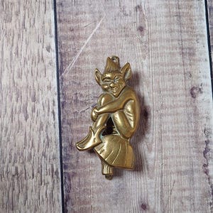 May include: A brass figurine of a seated goblin with a pointed hat and a mischievous grin. The goblin is depicted with its legs crossed and arms wrapped around its knees. The figurine has a vintage aesthetic.