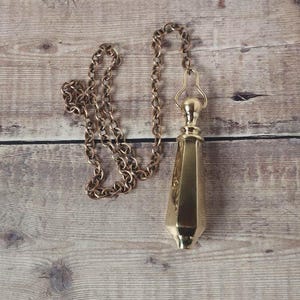 Könnte beinhalten: Eine goldfarbene Toiletten-Zugkette im Vintage-Stil mit einem dekorativen, facettierten Anhänger. Kette und Anhänger sind aus Metall und liegen auf einer Holzoberfläche. Der Anhänger hat eine strukturierte Oberfläche.