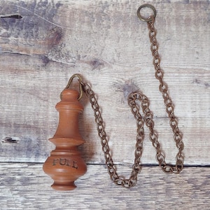 May include: A vintage wooden toilet pull with a chain. The pull is a light brown colour and has the word "FULL" engraved on it. The chain is made of metal and is a dark brown colour. The background is a wooden surface.