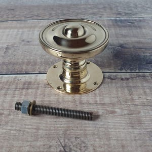 May include: A polished brass door knob with a rounded top and a circular base. A threaded bolt with a nut and washer is positioned beside the knob. The hardware is set against a wooden background.