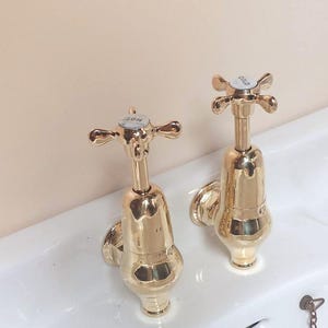 May include: Two polished gold-coloured taps with cross-shaped handles are fixed to a white sink. One tap is marked "HOT" and the other "COLD". The sink has the text "Armitage Ware" on it.