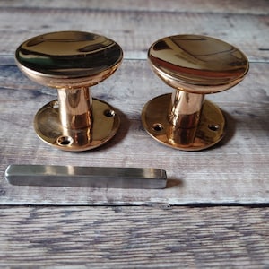 May include: Two polished brass door knobs with a round, mushroom-shaped top and a circular base. The knobs are on a wooden surface.