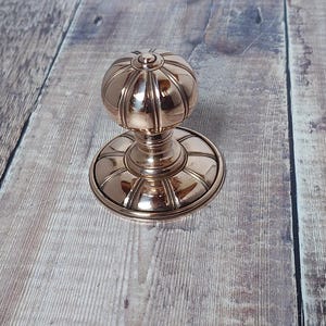 以下が含まれることがあります： 丸みを帯びたリブ付きのトップと円形のベースを備えた、ブロンズ色のドアノブ。 金具は金属製で、木製の表面に置かれています。 ドアノブはクラシックなデザインです。