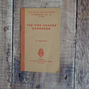 May include: A vintage handbook titled "The Fire Guards Handbook" with the text "Air Raid Precautions Handbook No. 14 (1st Edition)". The cover is tan with red text and a red spine. The book is on a wooden surface.