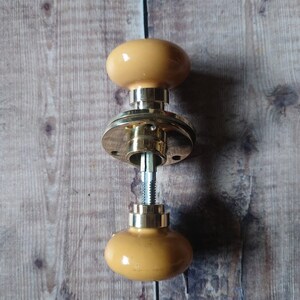 May include: A brass door knob with a yellow ceramic knob on each side. The knob is disassembled and the parts are laying on a wooden surface.