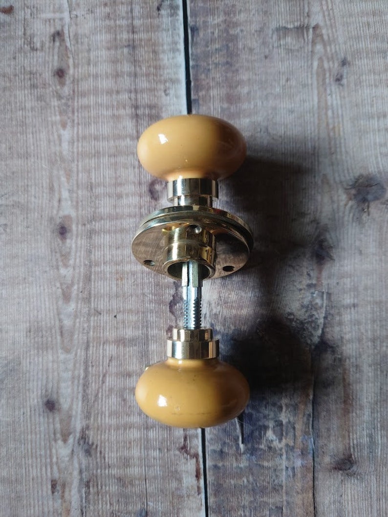 May include: A brass door knob with a yellow ceramic knob on each side. The knob is disassembled and the parts are laying on a wooden surface.