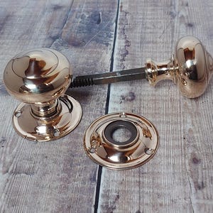 May include: A set of polished brass door knobs and a matching backplate. The knobs are round and shiny, connected by a metal spindle. The backplate is circular with screw holes, all on a wooden surface.