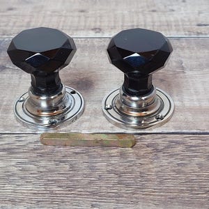 Könnte beinhalten: Zwei schwarze, facettierte Türknöpfe aus Glas mit silberfarbenen Metallsockeln und einer Metallverbindungsstange. Die Knöpfe haben ein geometrisches Design und sind vor einem Holzgrund platziert.