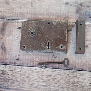 Könnte beinhalten: Ein antikes Schloss aus Metall für eine Tür mit einem Schlüsselloch und einem Schlüssel. Das Schloss ist rostig und hat eine braune Patina. Der Schlüssel ist silberfarben und hat ein schlichtes Design.