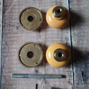 May include: A set of two brass door knobs with a yellow ceramic finish. The set includes two knobs, two backplates, a screw, an allen wrench, and two decorative pieces.