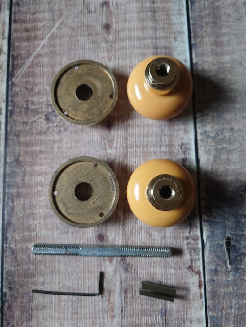 May include: A set of two brass door knobs with a yellow ceramic finish. The set includes two knobs, two backplates, a screw, an allen wrench, and two decorative pieces.
