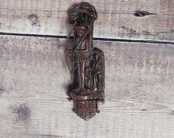 May include: A bronze-colored metal door knocker featuring a figure of Robin Hood with a bow and arrow. The knocker has a shield at the bottom with a coat of arms and the words "Robin Hood" are visible.