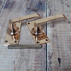 以下が含まれることがあります： 正方形のバックプレートが付いた、磨かれた銅製のドアノブが2つ。ノブは木製の表面の上に置かれています。