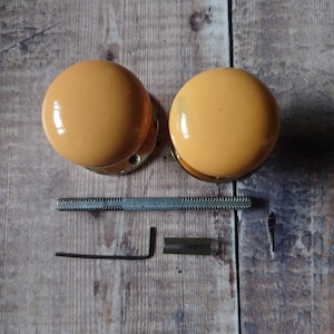 May include: Two round, yellow ceramic door knobs with gold metal hardware. The knobs are on a wooden surface with a metal screw, allen wrench, and a small metal piece.