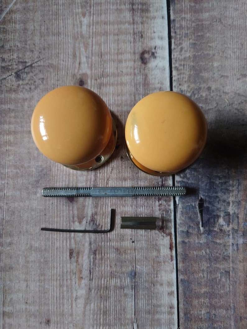 May include: Two round, yellow ceramic door knobs with gold metal hardware. The knobs are on a wooden surface with a metal screw, allen wrench, and a small metal piece.