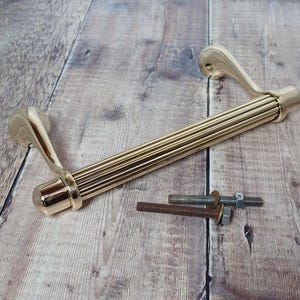 May include: A gold-coloured door handle with a ribbed central section and curved ends. Two bolts with washers and nuts are next to the handle. The handle is on a wooden surface.