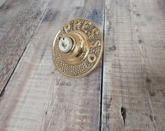 May include: A vintage brass doorbell button with the word "PRESS" embossed on the circular face. The button has a central white ceramic insert with a small illustration. The button is mounted on a wooden surface.