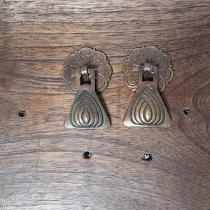 May include: Two antique brass drawer pulls with a floral design and a triangular handle. The pulls are mounted on a dark wood surface. The hardware is likely for a cabinet or dresser.