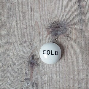 May include: A white, round ceramic knob with the word "COLD" printed in black. The knob is set against a weathered, light brown wooden surface, showcasing a rustic aesthetic.
