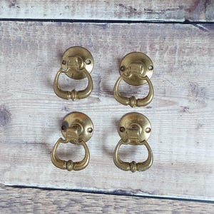 May include: Four antique brass drawer pulls with ring handles. Each pull has a circular backplate with a curved handle. The pulls are arranged in a square pattern on a wooden surface.
