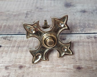 May include: A brass-toned, cross-shaped door knob with a central circular design and four ornate, star-shaped arms. The knob has a vintage aesthetic and is set against a wooden background.