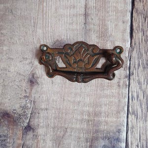 May include: An ornate bronze drawer pull with a decorative floral design. The handle has a curved shape and is mounted on a wooden surface. The hardware is likely vintage or antique.