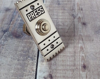 May include: A rectangular, silver-colored doorbell with the word "PRESS" in a black, rectangular box. The doorbell has a circular button and decorative lines and triangles along the edges. The doorbell is mounted on a wooden surface.