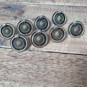 May include: A collection of eight antique-style drawer pulls. Each pull features a circular design with a central floral pattern and a ring handle. The pulls have a silver and bronze finish, and are arranged on a wooden surface.